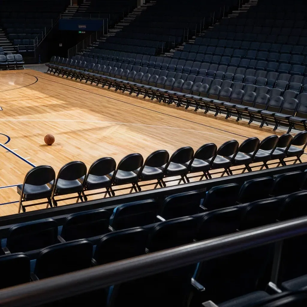 Empty basketball arena with a ball near center court and folded chairs under warm late-afternoon light.